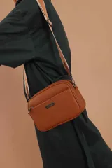 Bandolera de diseño rectangular con textura granulada, cuenta con tres compartimentos principales con cierre, un bolsillo frontal y un bolsillo trasero. Incluye una correa larga ajustable y extraíble con detalles en contraste, y una placa metálica decorativa en el frente.