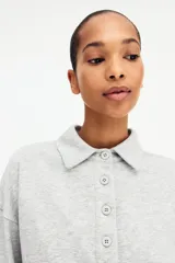 Buzo en tejido liviano color gris melange, con cuello camisero, tapeta con botones frontales, hombros caídos y puños acanalados.