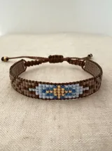Pulsera tejida con cuentas Miyuki, con diseño geométrico en tonos marrón, celeste, azul y dorado. Cuenta con un sistema de cierre ajustable mediante cordón marrón.