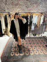 Ruana tejida a mano en lana merino color beige, con textura de punto y detalle de flecos en el ruedo. Presenta sutiles destellos brillantes integrados en el tejido.