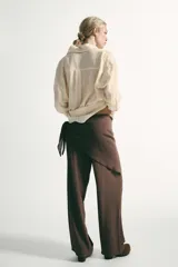 Pantalón de tiro medio con pernera recta y amplia, confeccionado en tejido con textura. Presenta una pieza superpuesta tipo pañuelo anudada en la cintura y bajo ajustable con botón.