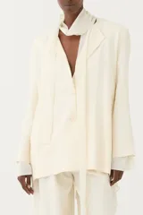 Blazer oversize de color blanco marfil, con solapas, cierre frontal de dos botones y mangas largas. Incluye un pañuelo integrado de la misma tela para anudar al cuello.