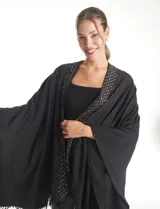 Ruana de tejido soft color negro, con detalles de brillos y tachas en los bordes frontales. Presenta una caída fluida y terminación con flecos.