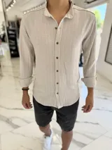 Camisa de lino color tostado con sutiles rayas verticales blancas, cuello mao y mangas largas con puños vueltos. Cuenta con cierre frontal de botones oscuros.