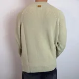 Sweater de tejido de punto grueso color verde militar, con cuello redondo y mangas largas tipo raglán. Presenta un corte holgado y una pequeña etiqueta con logo en el puño.