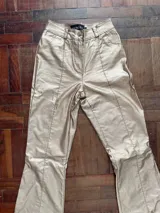 Pantalón de tiro alto confeccionado en eco cuero con acabado metalizado dorado. Presenta un corte recto con aberturas laterales con cierre en los tobillos y costuras frontales decorativas.