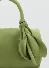 Bolso de mano cilíndrico de cuero gamuzado color verde, con dos asas cortas y detalle de lazo decorativo en un lateral. Cuenta con cierre superior de cremallera.