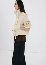 Chaqueta crop estilo gabardina corta color beige, de diseño recto, cuello alto tipo embudo, manga larga, bolsillos laterales y cierre cruzado de doble botonadura con botones oscuros. Con forro interior de poliéster.