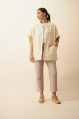 Chaleco recto sin mangas, color crema/beige claro, con cuello y solapas pronunciadas, cierre frontal con broches a presión y bolsillos ojal. Presenta terminaciones envivadas a la vista.