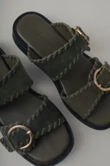 Sandalia de plataforma color verde militar con doble tira ancha. Las tiras tienen un acabado de gamuza con pespuntes decorativos y hebillas metálicas doradas. La suela es gruesa y negra.