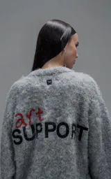 Sweater de punto texturizado color gris, con cuello redondo y diseño oversize. Presenta un bordado en relieve en la espalda con la frase 'art SUPPORT' en letras rojas y negras.