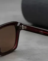 Lentes de sol unisex modelo Adisa, con montura gruesa de forma cuadrada y patillas robustas. El color de la montura es marrón oscuro (maroon) y los lentes son de color marrón.