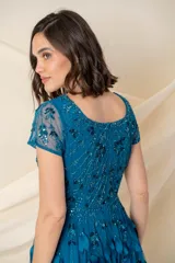 Vestido largo de tul color petróleo, con escote redondo y mangas cortas. Presenta un cuerpo bordado con lentejuelas al tono y una falda con movimiento decorada con apliques de hojas de tela.