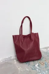 Bolso tipo tote de cuero vacuno con textura granulada, color bordó. Presenta doble asa de hombro y un detalle de lazo decorativo en la parte superior central.