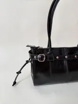 Cartera de cuero de tamaño medio con estructura rígida. Presenta una tira decorativa con tachas y hebillas metálicas en los extremos, además de lazos laterales. Cuenta con doble asa para llevar al hombro y cierre superior.