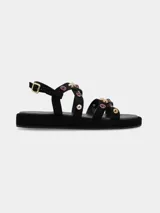 Sandalias planas de cuero gamuza en color negro, con múltiples tiras cruzadas decoradas con tachas metálicas doradas y aplicaciones de piedras rojizas. Cuenta con suela de PVC y tira ajustable en el talón con hebilla metálica dorada.