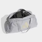 Bolso deportivo Adidas de color gris claro con logo estampado en tono beige. Cuenta con compartimento principal con cierre, bolsillo frontal con cremallera, correas de mano y correa de hombro ajustable con hombrera acolchada.