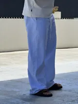Pantalón de corte ancho y holgado, con diseño de rayas verticales finas en color azul y blanco. Presenta cintura elástica con cordón ajustable y un pequeño bordado en color rojo en la parte superior.