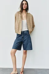 Chaqueta bomber de color beige con diseño a cuadros blancos, cuello subido, manga larga con trabillas en los hombros, bolsillos delanteros y bajo con elástico. Cierre frontal con cremallera y botones a presión.