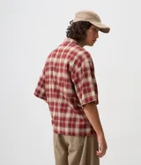 Camisa de franela de algodón con diseño a cuadros en tonos rojo y beige, de corte holgado relaxed, mangas cortas, cuello tradicional y cierre frontal con cremallera. Cuenta con un bolsillo aplicado en el pecho con detalle de botón metálico.