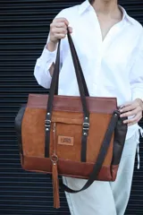 Bolso tipo shopper de Lorena Caprile, confeccionado en una combinación de cuero vacuno graneado y gamuza en tonos marrón chocolate, óxido y whisky. Presenta doble asa de hombro, correa larga ajustable, bolsillo frontal con cierre, detalle de borla de cuero con aro metálico y logo de la marca grabado en el frente.