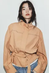 Chaqueta color tostado con cuello subido, manga larga y detalle de tachas metálicas en contraste. Incluye cinturón del mismo tejido con hebilla metálica y cierre frontal con cremallera y botones a presión ocultos.