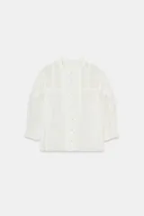 Camisa blanca de algodón con cuello redondo y manga por debajo del codo abullonada. Presenta detalles de bordados a tono y cierre frontal con botones.