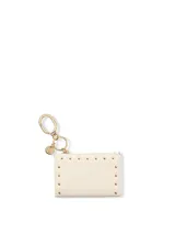 Estuche para tarjetas y llavero color marfil de cuero sintético, decorado con tachas doradas a lo largo de los bordes superiores y laterales. Incluye un sistema de argollas y gancho mosquetón metálico dorado con un dije redondo con logo grabado.