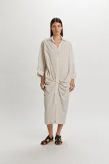 Vestido camisero midi color beige con sutiles rayas verticales oscuras, de manga larga, cuello camisero, cierre frontal con botones y detalle de fruncidos drapeados en los laterales de la falda.