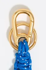 Llavero con mosquetón y anillas de metal dorado, que incluye un dije central en forma de corazón con el logo grabado. Presenta un cuerpo de cordones trenzados en color marrón con motas blancas y terminaciones anudadas.