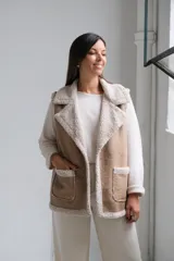 Chaleco de gamuza color beige con interior y detalles de corderito color crudo. Presenta solapas amplias, bolsillos frontales aplicados y diseño sin mangas.