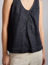 Top sin mangas de lino color negro con escote en V y detalle de bordado central en forma de trébol de cuatro hojas. Presenta costuras decorativas visibles en el escote y el ruedo.