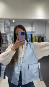 Chaleco corto de efecto metalizado en tono celeste claro, con cuello en V, cierre frontal con un botón a presión y dos bolsillos delanteros con solapa.