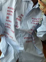 Camisa oversize de corte recto, confeccionada en tela de algodón a rayas verticales celestes y blancas. Presenta cuello clásico, cierre frontal con botones y mangas largas. El diseño destaca por una estampa frontal con palabras relacionadas al amor en diversos idiomas en color bordó.
