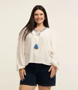 Blusa color blanco con escote en pico con bordado de corazones azules, mangas largas con puños elásticos, lazos con borlas y detalle de hilo metalizado.