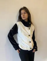 Campera corta de textura suave, con diseño bicolor en blanco y negro. Presenta un cierre frontal con botones dispuestos en diagonal, cuello alto envolvente y mangas largas en color negro.