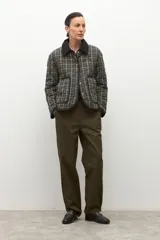 Campera capitoneada de corte recto con diseño a cuadros en tonos verde oliva y gris. Presenta cuello camisero de pana en color negro, cierre frontal con botones a presión y bolsillos laterales amplios con ribete negro.