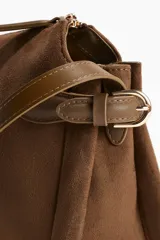 Cartera cruzada de material tipo gamuza color marrón oscuro, con cierre superior y correa ajustable delgada con hebilla metálica dorada. Presenta trabillas decorativas en los laterales.