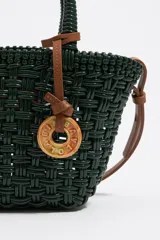 Bolso tipo cesta de estructura rígida y diseño trenzado en color negro. Cuenta con dos asas superiores cortas y una correa larga ajustable en color marrón. Incluye un dije circular decorativo con grabado en tono madera.