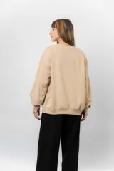 Buzo oversize de algodón frizado color beige, con cuello redondo, puños y cintura tejidos. Presenta un bordado pequeño de Snoopy en el frente.