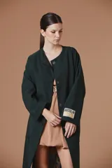 Saco Natalia Antolin de mujer color verde oscuro, de corte largo y diseño relajado con cuello redondo, mangas anchas y cierre frontal de botones oscuros. Incluye una etiqueta decorativa distintiva de la marca en el puño de la manga izquierda.