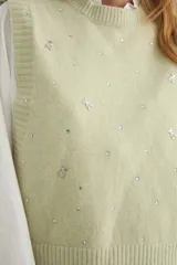 Chaleco de punto color verde salvia, con cuello redondo y diseño sin mangas. Presenta detalles de pedrería brillante y pequeños apliques decorativos distribuidos por toda la prenda.