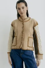 Chaqueta de punto en tonos beige y crema, con diseño de paneles combinados. Presenta mangas con tejido de ochos, cuerpo con costuras decorativas a la vista, cierre frontal con botones y bolsillos delanteros aplicados.