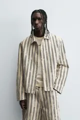 Cazadora de hombre de corte relajado, color beige claro con rayas verticales en azul oscuro con efecto desgastado o estampado. Presenta cuello solapa, manga larga, bolsillos de vivo en cadera y cierre frontal con botones a presión.