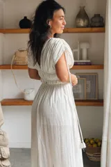 Vestido largo color blanco, con escote en V, mangas cortas abullonadas y cintura fruncida elastizada. Presenta detalles de encaje y cordones con borlas en el frente, además de una abertura lateral en la falda.
