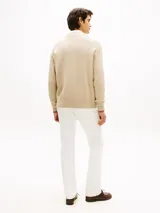 Cárdigan de punto grueso en color beige, con cuello en V, cierre frontal con botones y bolsillos de ojal. Presenta puños y bajo acanalados con líneas rojas y blancas, y un logo script bordado en rojo en el pecho.