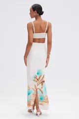 Vestido largo blanco con estampado tropical en tonos turquesa, naranja y marrón en el ruedo. Presenta escote corazón con detalle de nudo en el busto, recortes laterales en la cintura y tirantes finos con herrajes dorados.