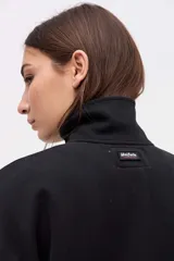 Buzo oversize de felpa con friza, color negro, con cuello alto y cierre frontal parcial metálico. Presenta bolsillos laterales, tajos en los costados y un diseño con espalda más larga. Incluye un pequeño logo bordado en el pecho.