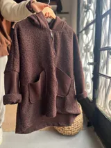 Campera oversize de textura tipo corderito en color marrón, con cierre frontal, capucha y bolsillos delanteros amplios. Presenta un diseño de corte holgado con mangas largas y terminaciones en contraste.
