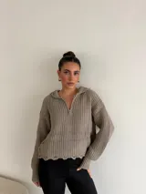 Buzo de punto grueso color gris, con cuello polo, cierre frontal parcial y capucha. Presenta un diseño de tejido acanalado con terminaciones desflecadas en el ruedo.